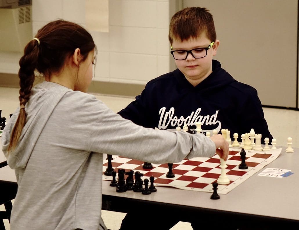 Chess Club picture of chess players. A girl is in the foreground making a move with her King which is on h2. A boy with glasses closely observes her move. He has a bishop on h6.