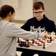 Chess Club picture of chess players. A girl is in the foreground making a move with her King which is on h2. A boy with glasses closely observes her move. He has a bishop on h6.