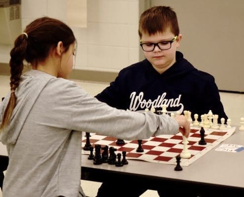 Chess Club picture of chess players. A girl is in the foreground making a move with her King which is on h2. A boy with glasses closely observes her move. He has a bishop on h6.